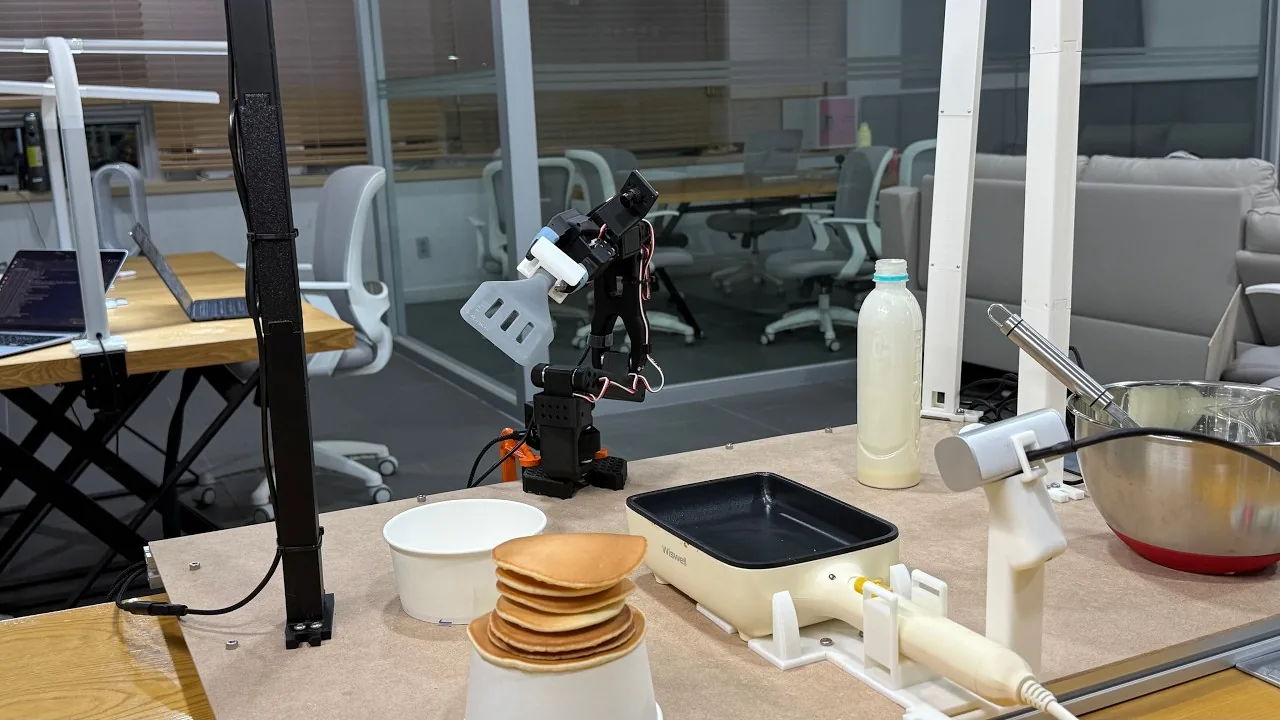 The Panbot workstation: SO-ARM101 arm with a 3D-printed spatula, electric griddle, batter dispenser, and a stack of finished pancakes